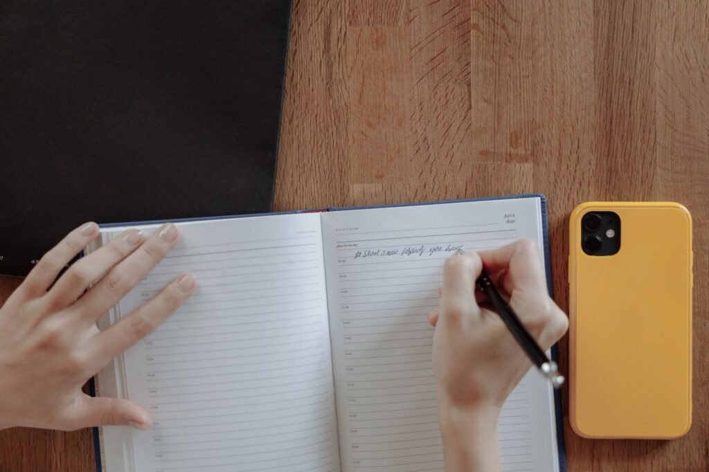 A person writing notes in a notebook beside a smartphone on a wooden table